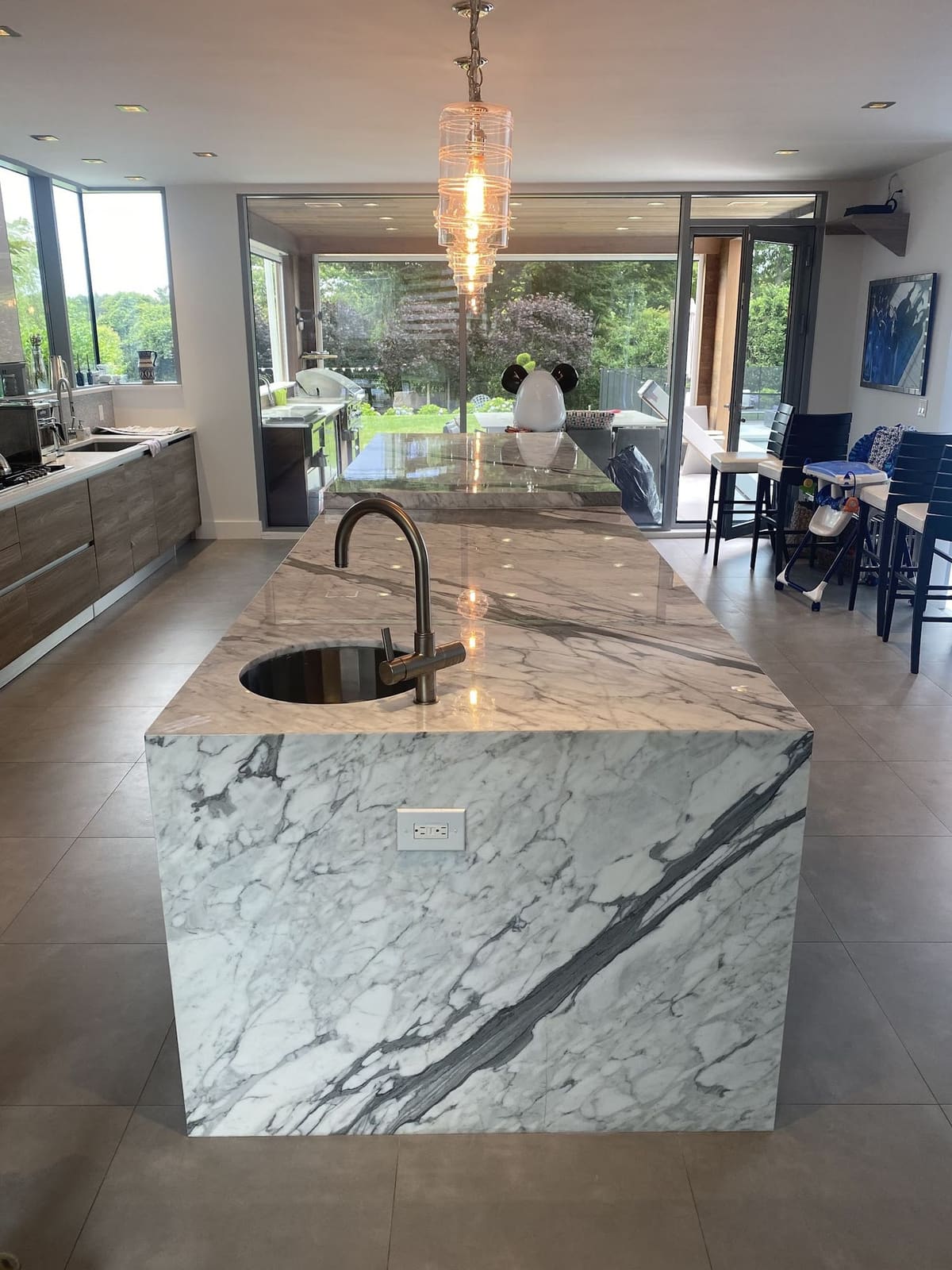 Kitchen island wrapped in white marble with bold gray veining under a crystal pendant, open to the lanai