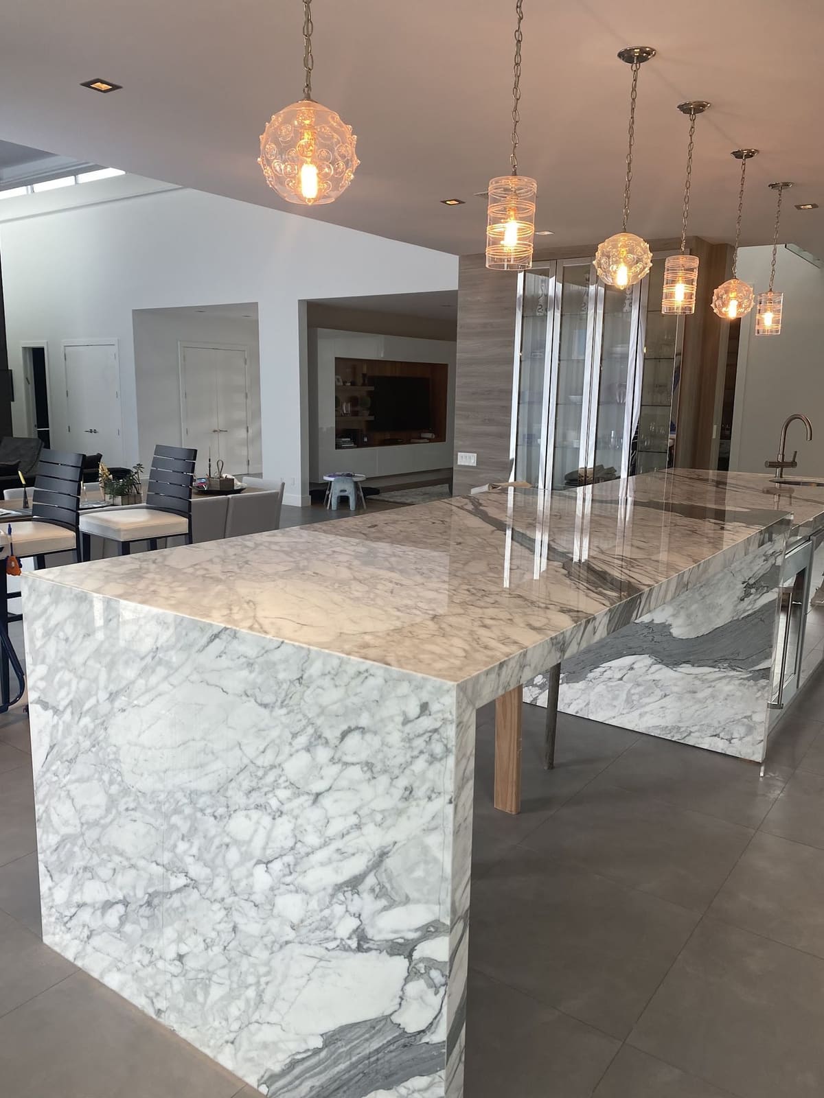 Long waterfall-edge marble island beneath a row of mixed glass pendant lights in an open-plan home